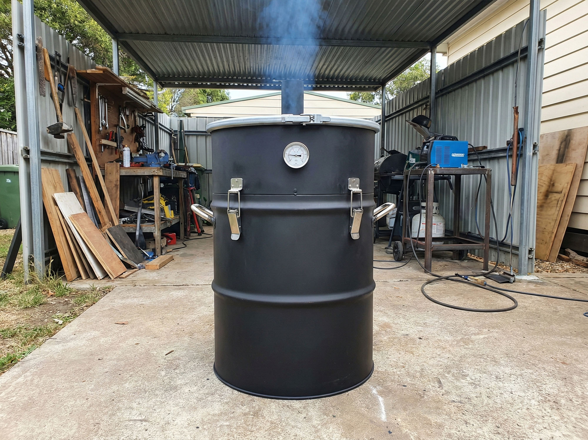 A custom-built black steel drum smoker with silver hardware in a backyard workshop setting.