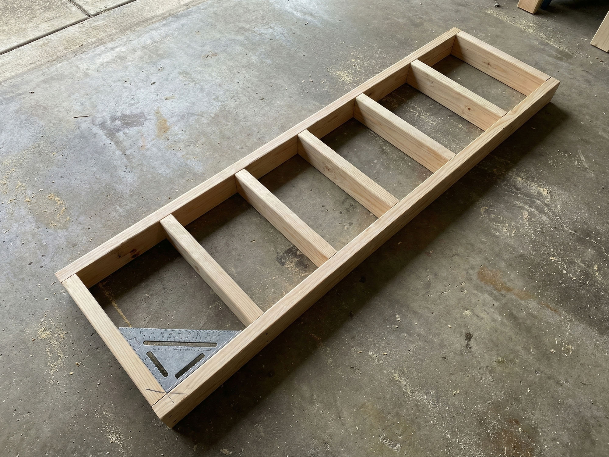 High-angle view of a completed wooden ladder frame for a workbench made from 2x4 lumber on a concrete garage floor with a speed square.