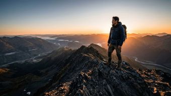 日の出の中、広い谷を見渡す岩山の頂上に立つ男性。